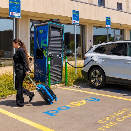 En vit elbil laddas vid en publik laddstation framför ett hotell.